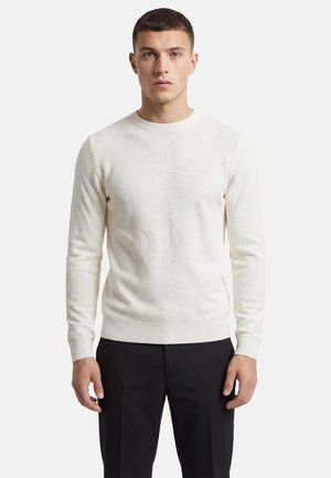 Ung mand stående, iført en tekstureret hvid sweater og sorte bukser, vendt fremad med et neutralt udtryk på en ensfarvet baggrund.