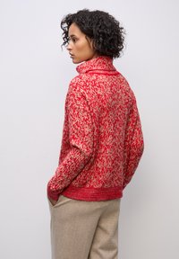 Suéter de punto con patrón rojo y crema, cuello alto, puños acanalados y un corte relajado, combinado con pantalones beige.