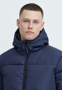 Jachetă puffer cu glugă, de culoare bleumarin, cu închidere prin fermoar, având o textură matlasată și un guler înalt pentru confort termic.