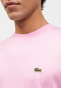 Κοντινό πλάνο ενός ατόμου που φοράει ανοιχτό ροζ πουκάμισο με ένα μικρό πράσινο κροκοδειλάκι κεντημένο στο στήθος.