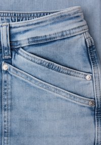 Jean en denim bleu clair avec poche avant visible, petite poche, passants de ceinture et détails de rivets argentés sur tissu texturé.