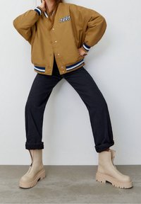 Osoba w beżowej kurtce studenckiej z naszywką "1991", czarnych spodni w rurki podwiniętych na dole oraz beżowych, masywnych butach na zamek, stoi z nogami rozstawionymi.