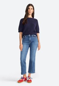 Pull en tricot navy à manches courtes, jeans droits bleus et baskets rouges avec lacets bleus et semelles beiges sur un fond gris clair.