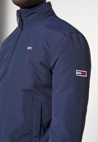 Granatowa kurtka z wysokim kołnierzem, przednim zamkiem błyskawicznym i małym logo w kolorach czerwonym, białym i niebieskim na lewym rękawie. Gładka tkanina, dopasowany krój.