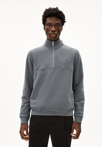 Felpa grigia a mezza zip realizzata in materiale morbido, con colletto alto, polsini a coste e un piccolo logo sul petto. Design semplice.