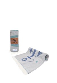 Rolled and partially unrolled light gray towel with blue anchor and fish designs, fringed edges, and a brown label band on the rolled towel.