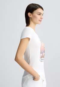 T-shirt blanc à manches courtes avec un design graphique coloré représentant une scène de plage. Texture lisse et coupe décontractée, avec un col rond.