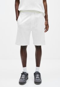 Shorts en coton blanc à mélange avec un taille élastique à cordon, poches latérales et une coupe décontractée. Associés avec des chaussures de sport noires et grises et des chaussettes blanches.