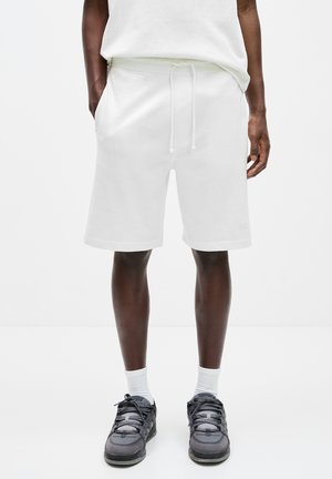 Weißer Baumwollmischung-Shorts mit einem Kordelzugbund, seitlichen Taschen und einer entspannten Passform. Kombiniert mit schwarzen und grauen Sportschuhen und weißen Socken.