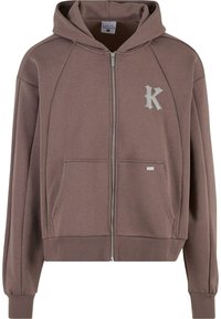 Felpa con zip color marrone, dotata di cappuccio, polsini e orlo a costine, grandi tasche frontali e un grande logo "K" argentato sul lato sinistro del petto. Materiale morbido.