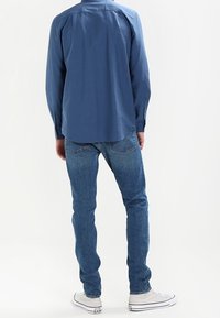 Hellblaues langärmliges Hemd aus Baumwollstoff, lockere Passform, mit Yoke-Detail auf der Rückseite. Trägt man mit blauen Jeans und weißen Turnschuhen.
