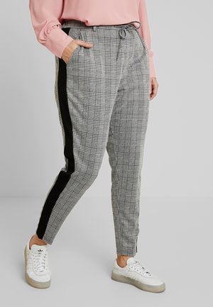 Person trägt graue karierte, schmal zulaufende Hosen mit schwarzen Seitenstreifen, ein rosa Langarmshirt und weiße Turnschuhe, steht mit einer Hand in der Tasche.