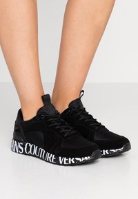 Svarta sneakers med en kombination av mocka och tyg, med en helt svart ovandel, gummisula och framträdande vit branding på sulan.