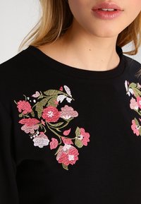 Sudadera negra con bordados florales coloridos en rosa, blanco y verde, que presenta un cuello acanalado y una textura suave.
