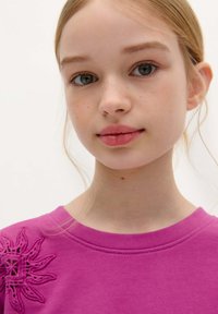 Rosa sweatshirt med en struktureret blomsterbroderi detalje på venstre skulder, rund hals og blødt stof, båret af en ung person.