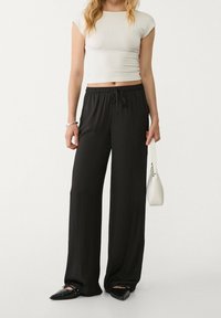 Žena v bílém crop topu, černých širokých kalhotách, černých špičatých polobotkách, s náramkem a držící malou bílou kabelku.