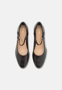 Scarpe in pelle nera con punta rotonda, cinturino alla caviglia e un piccolo dettaglio a fibbia. L'interno presenta un leggero padding con motivi perforati.