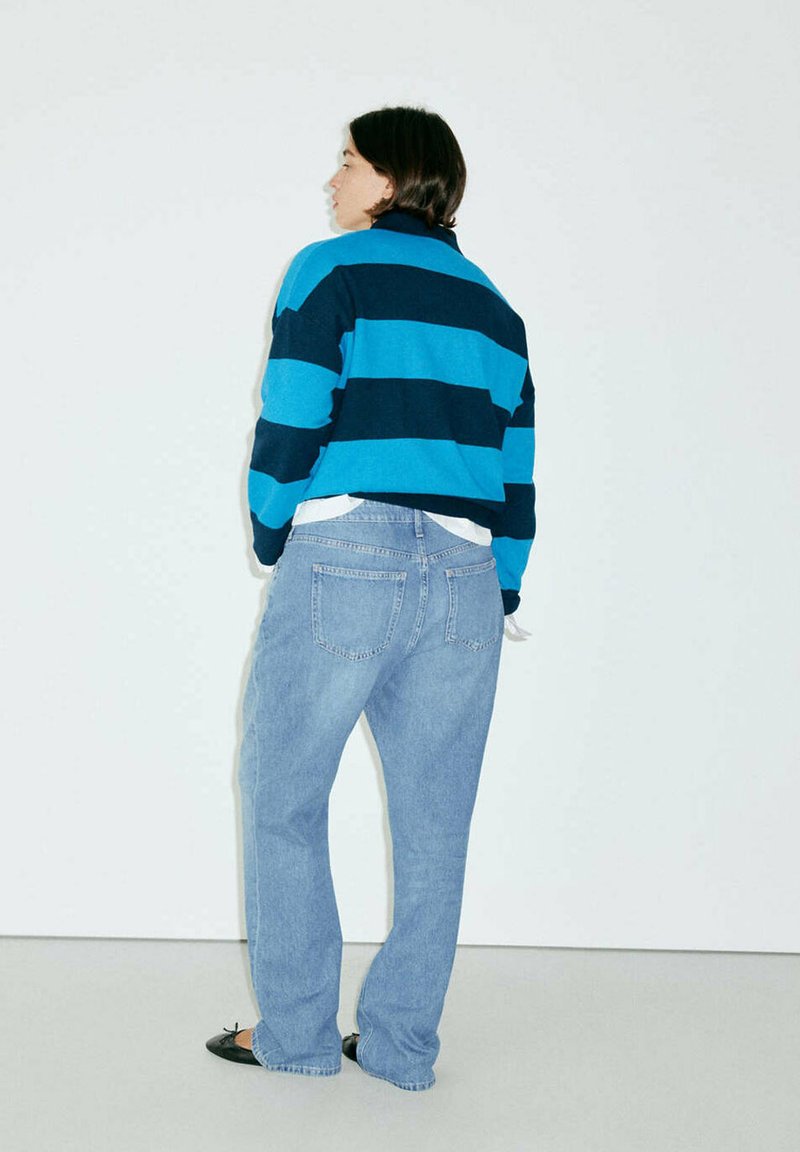 Personne debout face à un mur portant un pull rayé bleu et bleu foncé, un jean bleu clair et des chaussures plates noires.