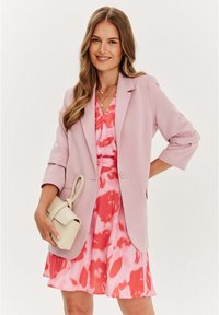 Světle růžový blejzr s rukávy po třičtvrtě přes růžové šaty s akvarelovým vzorem v odstínech růžové a červené. Drží krémovou kabelku.