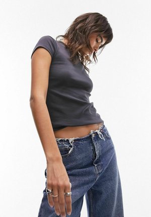 Γυναίκα που φοράει εφαρμοστή σκούρα γκρίζα μπλούζα και φαρδιά τζιν με ψηλή μέση και ξεφτισμένη μέση, κοιτάζει προς τα κάτω σε λευκό φόντο.