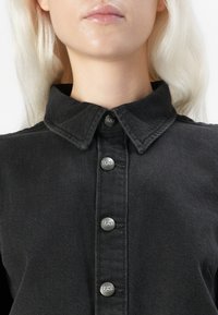 Sorte denimskjorte med spiss krage, sølvfarget knappelukking med "Lee"-merke og en strukturert overflate.