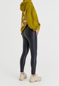 Leggings noirs ajustés associés à un sweat à capuche ample vert olive avec un texte blanc au dos. Le modèle porte des baskets de couleur claire.