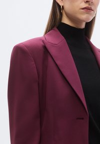 Bordeauxrode, op maat gemaakte blazer met scherpe revers en een enkele voorzak, gecombineerd met een zwarte top met hoge hals. Glad stof met gestructureerde pasvorm.