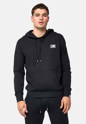 Jeune homme portant un sweat à capuche noir Leone 1947 et un pantalon de jogging noir assorti, debout devant un fond clair uni.