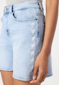 Bermudas de mezclilla azul claro con un diseño ajustado. Presenta corazones plateados bordados a lo largo de la costura lateral. Incluye un estilo estándar de cuatro bolsillos.