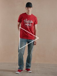 Tricou roșu din bumbac cu grafică "Vintage", blugi albaștri deschis și adidași roșii. Modelul poartă o șapcă neagră; fundal minimalist.