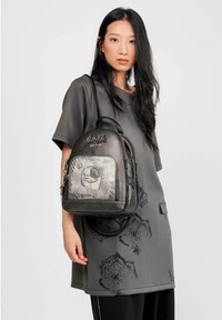 Mochila negra y gris con una superficie texturizada, que cuenta con un bolsillo frontal con cremallera y un diseño bordado. Modelo vistiendo un vestido gris.