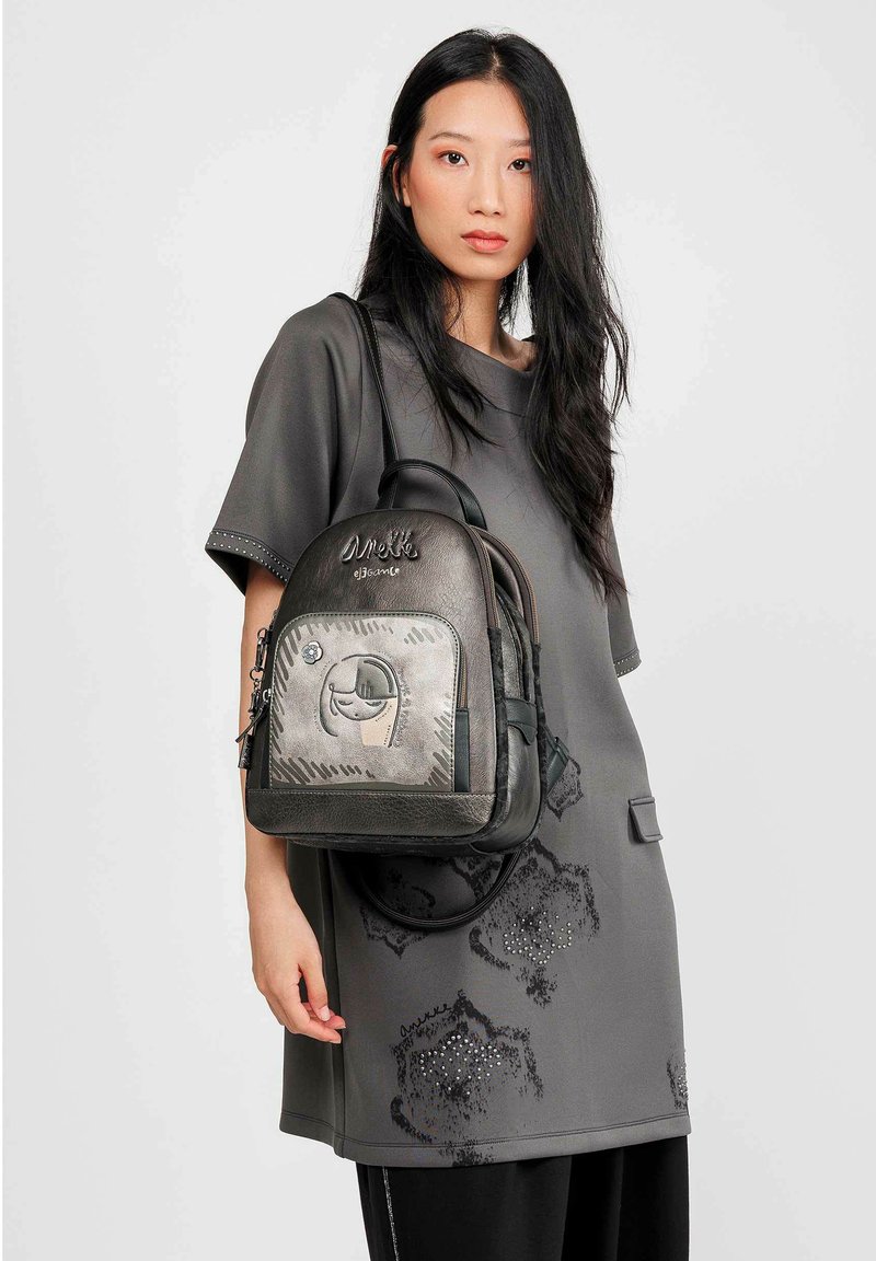 Mochila negra y gris con una superficie texturizada, que cuenta con un bolsillo frontal con cremallera y un diseño bordado. Modelo vistiendo un vestido gris.