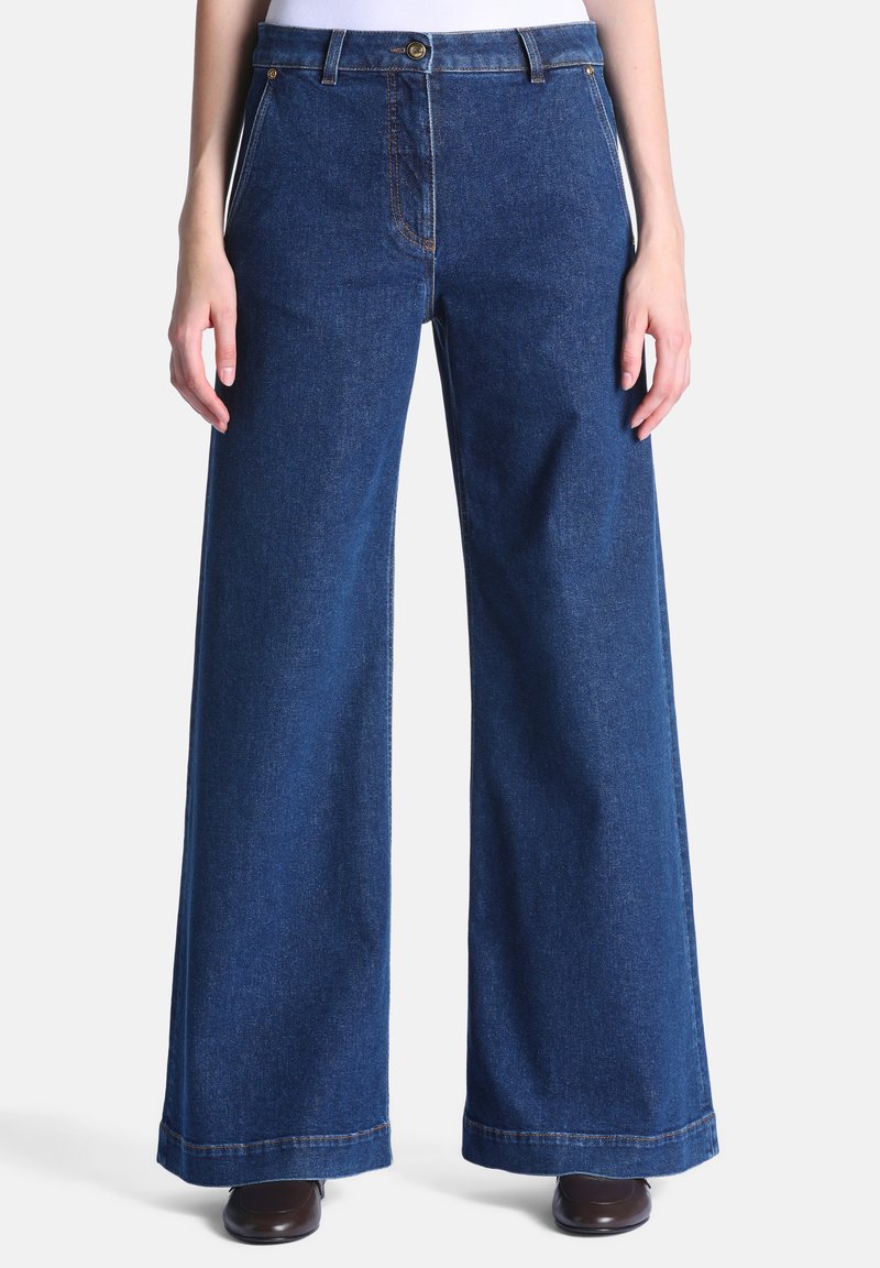 Jean en denim bleu à jambes larges, porté avec des chaussures foncées et un haut blanc rentré, vu de la taille jusqu’aux pieds, debout sur un fond blanc.