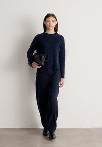 Γυναίκα με ίσια σκούρα μαλλιά, ντυμένη με μπλε πουλόβερ και παντελόνι με φαρδιά μπατζάκια, κρατώντας μαύρο τσαντάκι, στέκεται μπροστά σε έναν απλό λευκό τοίχο.