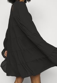 Robe noire à volants en tissu léger avec manches longues et coupe décontractée, présentant des volants subtils le long des coutures.