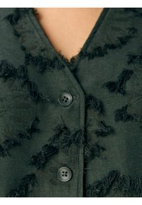 Gilet noir et vert en tissu texturé, avec des accents effilochés et deux boutons noirs à l'avant. Design en V.