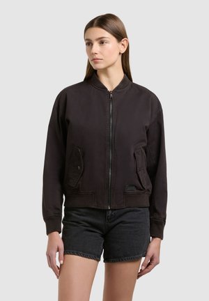 Kobieta ubrana w ciemnobrązową bomberkę na zamek z klapowymi kieszeniami oraz czarne krótkie spodenki jeansowe, stojąca na tle jednolitego szarego tła.