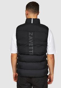 Zwarte puffer vest met een doorgestikt ontwerp, reflecterende ritssluiting aan de voorkant, en "ZAVETTI" branding in zilveren letters op de achterkant. Effen wit T-shirt eronder.