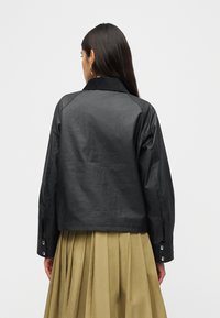 Γυναίκα με μακριά σκούρα μαλλιά που φοράει μαύρο cropped σακάκι με μανσέτες με κουμπιά και φούστα λαδί με πιέτες, εμφανιζόμενη από πίσω.