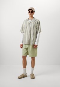Chemise à rayures à manches courtes en vert et blanc, associée à un short texturé vert clair. Les accessoires incluent une casquette beige et des lunettes de soleil.