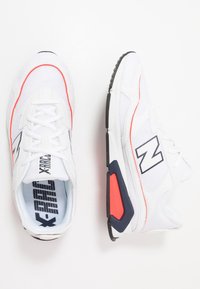 Bílé sportovní boty s prodyšným materiálem, s červeným akcentem a černou podrážkou. Obsahují tkaničky, vzorovanou stélku a design loga.