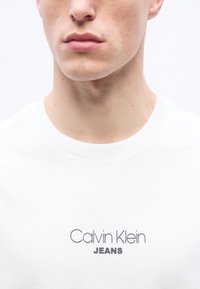 Κοντινό πλάνο του κάτω μέρους του προσώπου και του λαιμού ενός ατόμου που φοράει λευκό μπλουζάκι με το κείμενο "Calvin Klein JEANS" τυπωμένο στο στήθος.