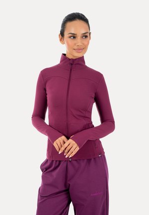 Kobieta ubrana w dopasowaną, zapinaną na zamek kurtkę z długim rękawem i luźne spodnie dopasowanego koloru śliwkowego, stoi ze splecionymi rękami, patrząc w prawo.