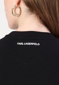 Gros plan du cou et des cheveux d'une personne portant une chemise noire avec « KARL LAGERFELD » imprimé sur le col arrière et une boucle d’oreille anneau dorée.