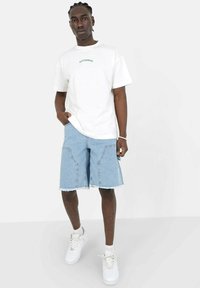 T-shirt bianco in cotone con testo verde, abbinato a pantaloni denim azzurri chiari con orli sfrangiati e grandi tasche frontali. Sneakers bianche.