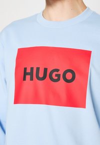 Sweatshirt bleu clair avec un grand graphisme rectangulaire rouge portant "HUGO" en lettres noires épaisses. Confectionné en tissu doux avec un col côtelé.
