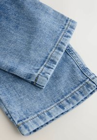 Haalari sinisen kuluneen farkkukankaan, jossa on sileä pinta ja käännetty helma. Reunoilla on keltaisia ompeleita.