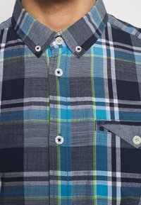 Rutig kortärmad skjorta i marinblått, blått och grönt, med knappkrage, fickdetalj och vita knappar.