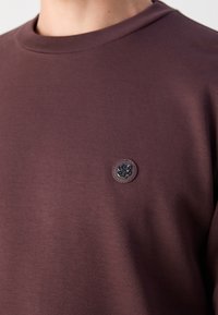 Sudadera de algodón marrón con cuello redondo y un parche de logo circular texturizado en el lado izquierdo del pecho. Tejido suave con un ligero brillo.