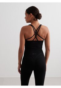 Juodas sportinis trumpas marškinėlis su kryžminėmis petnešomis, derinamas su juodomis aukštos juosmens legginėmis, turinčiomis tekstūruotą juosmenį ir logotipo detalę.